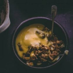 Spelt crumble in a dark bowl with thick custard