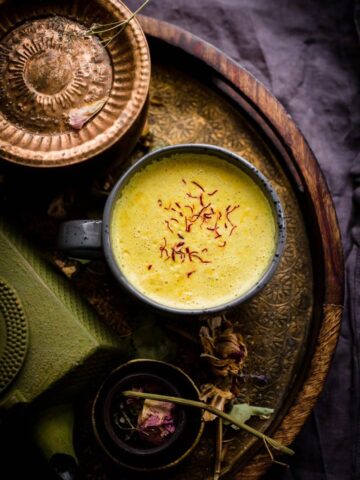 Turmeric and saffron milk in a cup, on gold tray, next to a green cast iron kettle