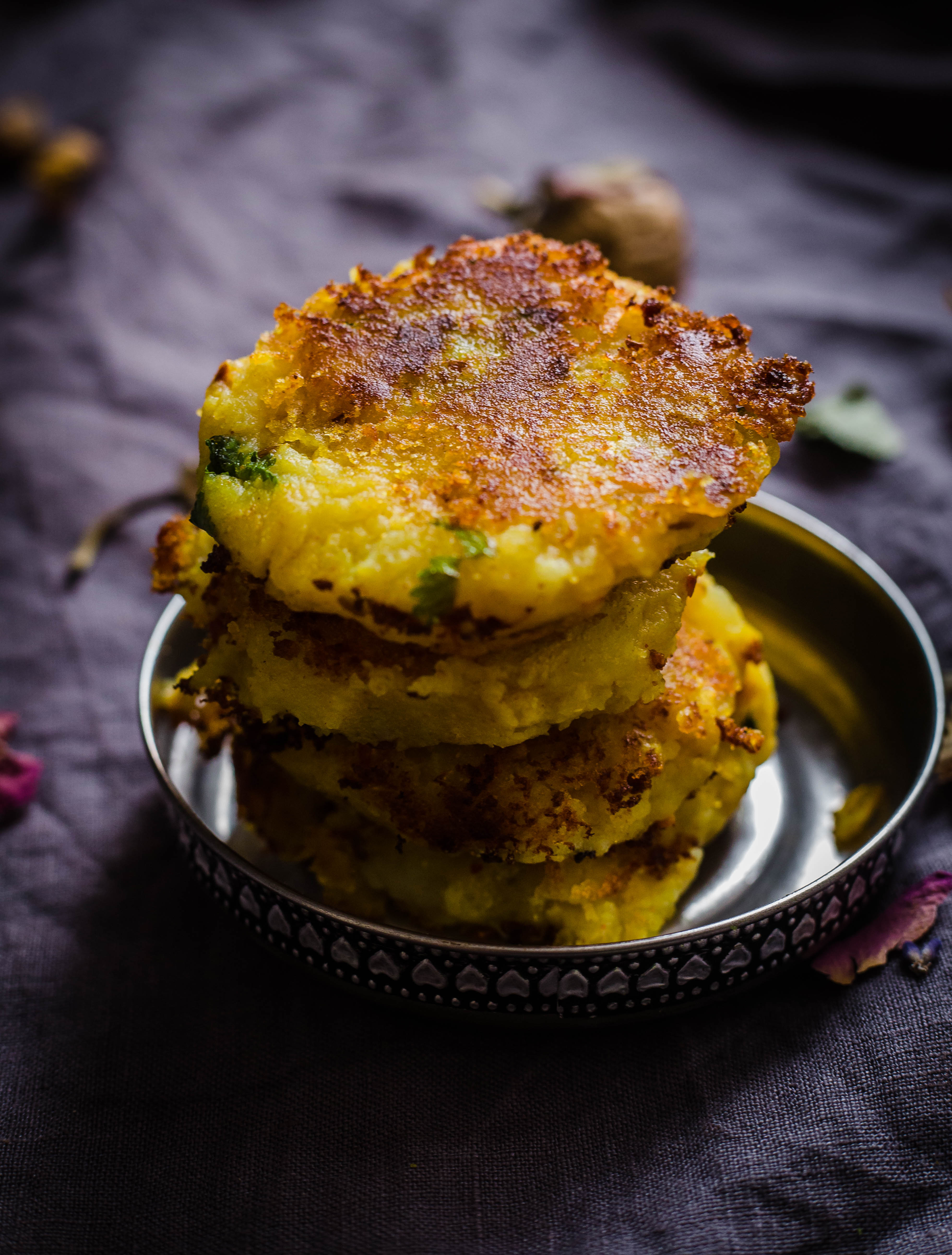 4 Aloo Tikkis on top of each other in a small silver lid on a grey towel with scattered dried flowers