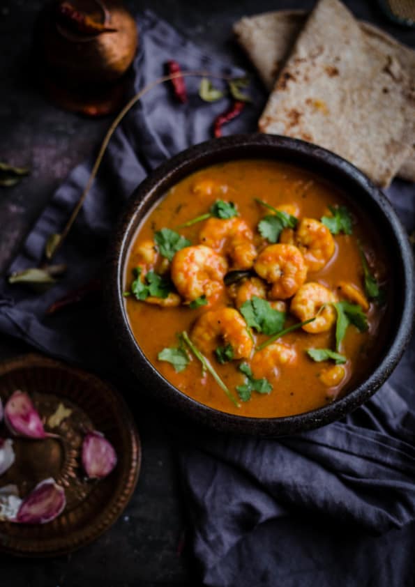 Prawn curry in bowl with roti to side and garlic cloves and curry leaves around 