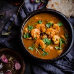 Prawn curry in bowl with roti to side and garlic cloves and curry leaves around