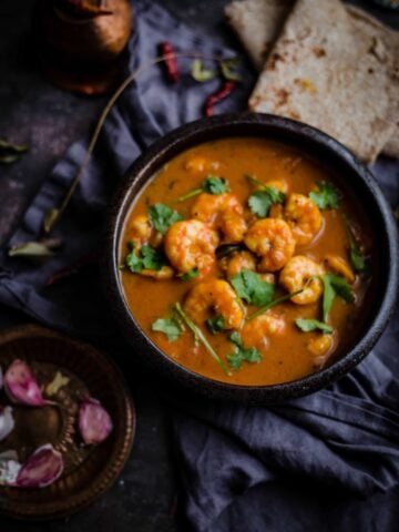 Prawn curry in bowl with roti to side and garlic cloves and curry leaves around